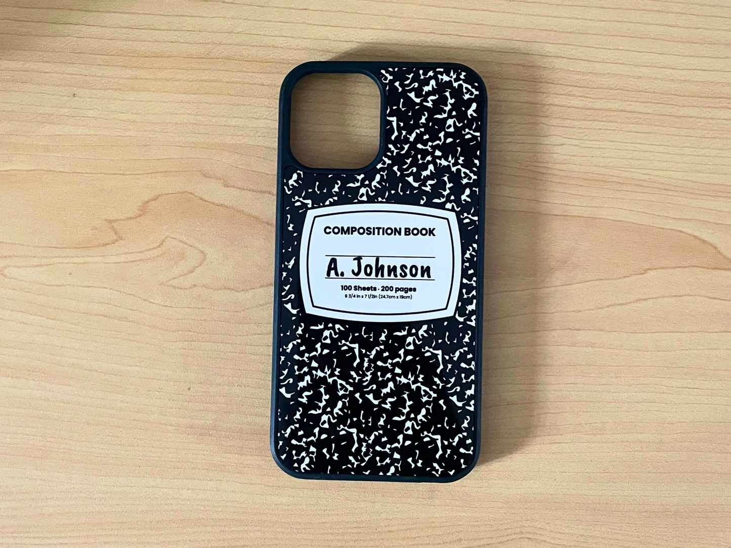 iPhone case with black and white composition notebook design and 'A. Johnson' written in the middle displayed on a wooden surface.