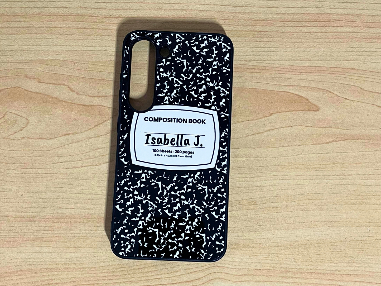 Samsung Galaxy case with black and white composition notebook design and 'Isabella J.' written in the middle displayed on a wooden surface.