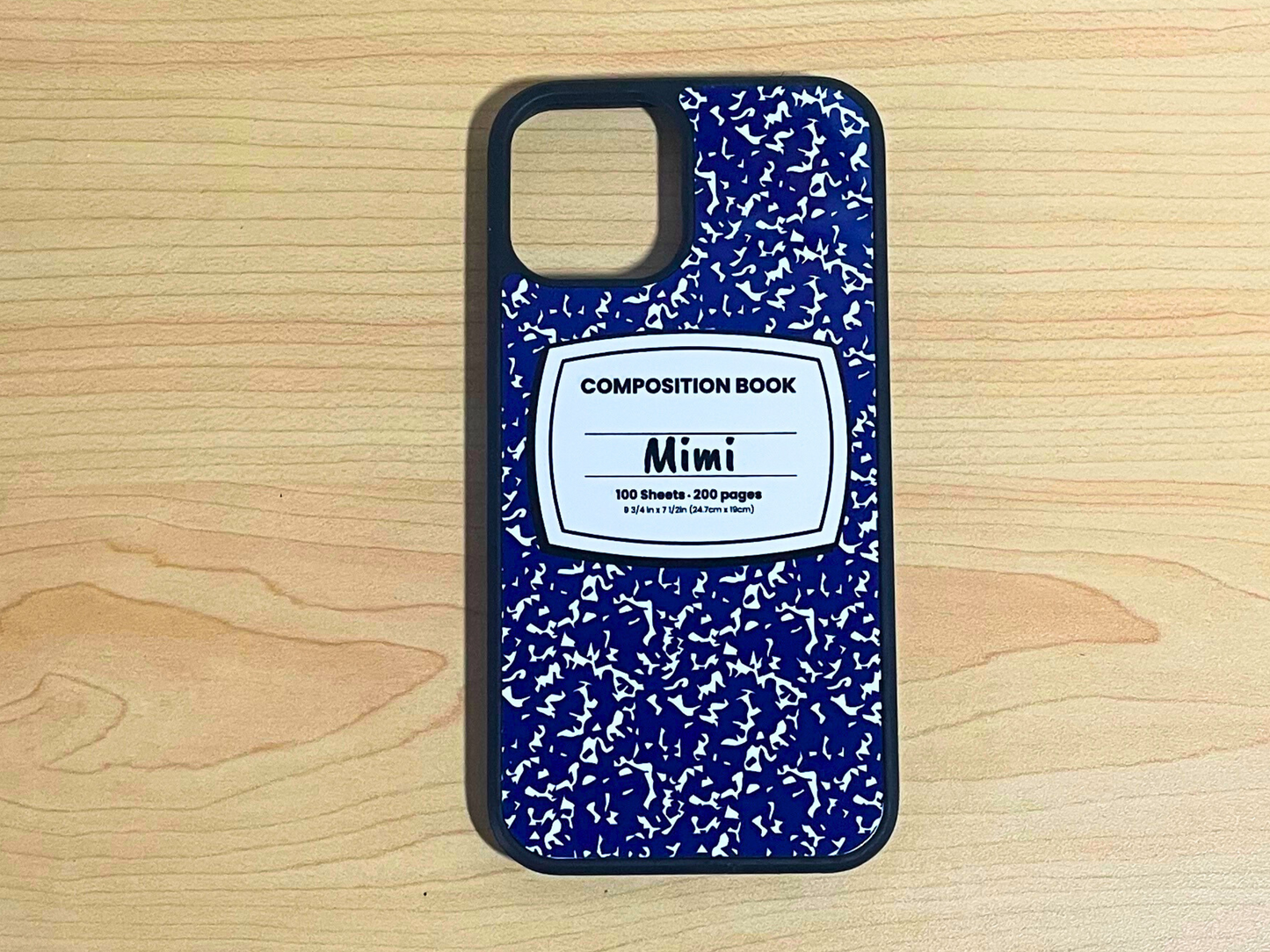 iPhone case with blue and white composition notebook design and 'Mimi' written in the middle displayed on a wooden surface.