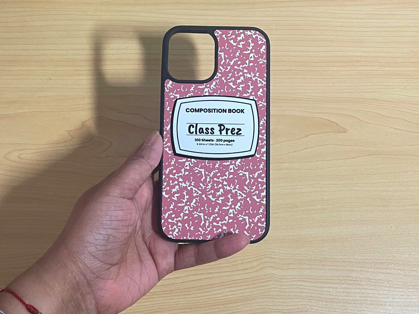 iPhone case with pink and white composition notebook design and 'Class Prez' written in the middle displayed on a wooden surface.