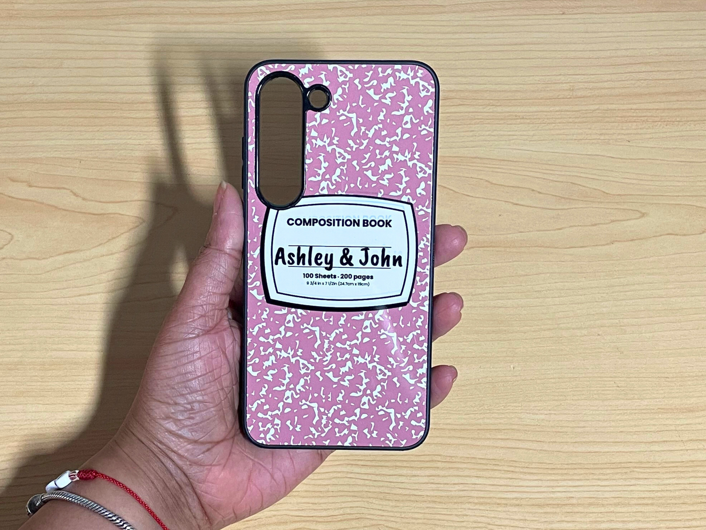 Samsung Galaxy case with pink and white composition notebook design and 'Ashley & John' written in the middle displayed on a wooden surface.