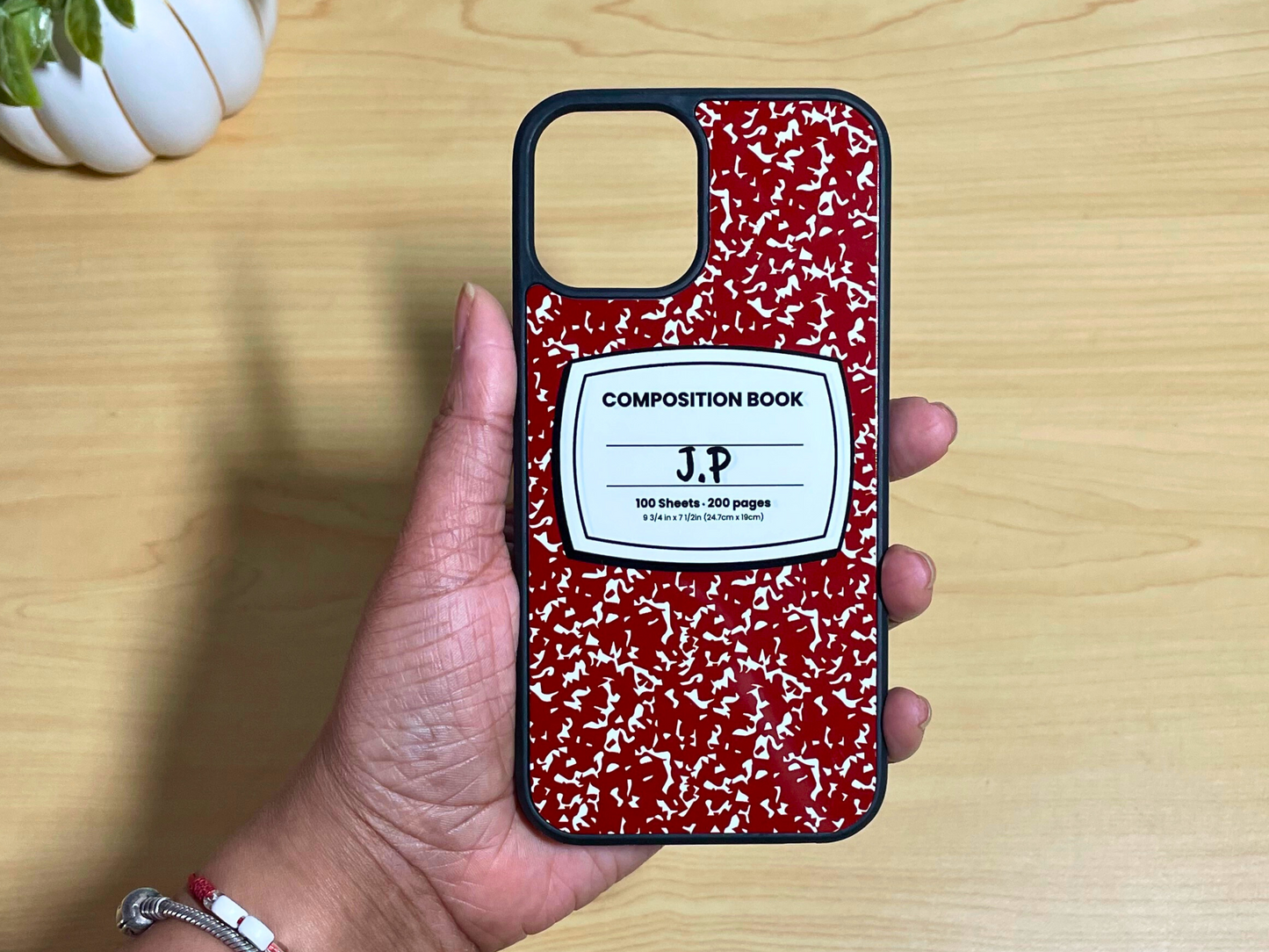 iPhone case with red and white composition notebook design and 'J.P' written in the middle displayed on a wooden surface.