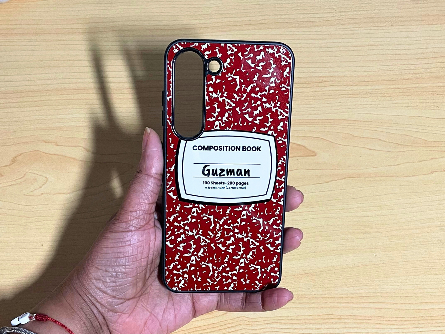 Samsung Galaxy case with red and white composition notebook design and 'Guzman' written in the middle displayed on a wooden surface.