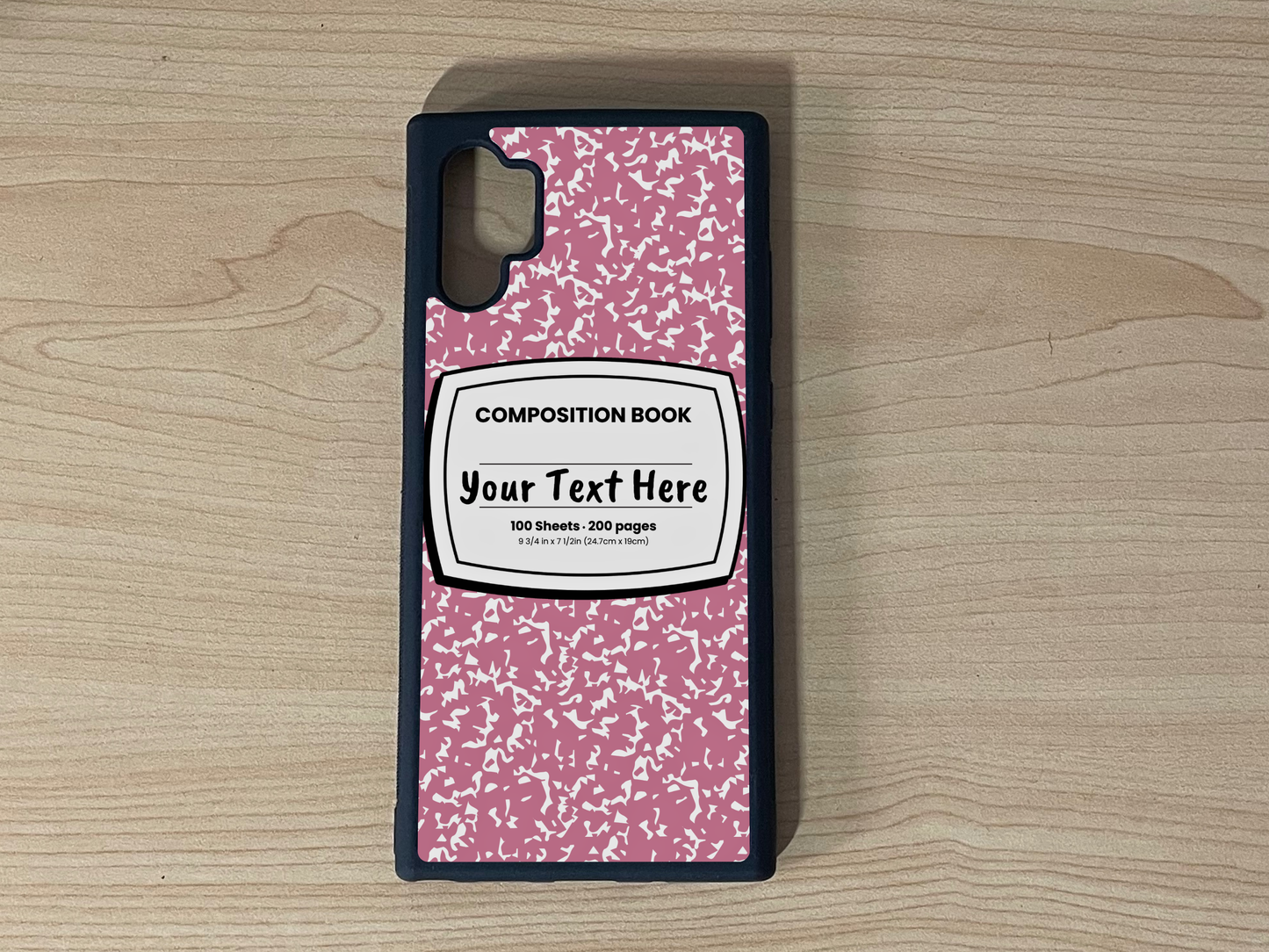 Custom phone case for the Samsung Note 10 Plus with a pink and white composition notebook design with text "Your text here" on a wooden surface.