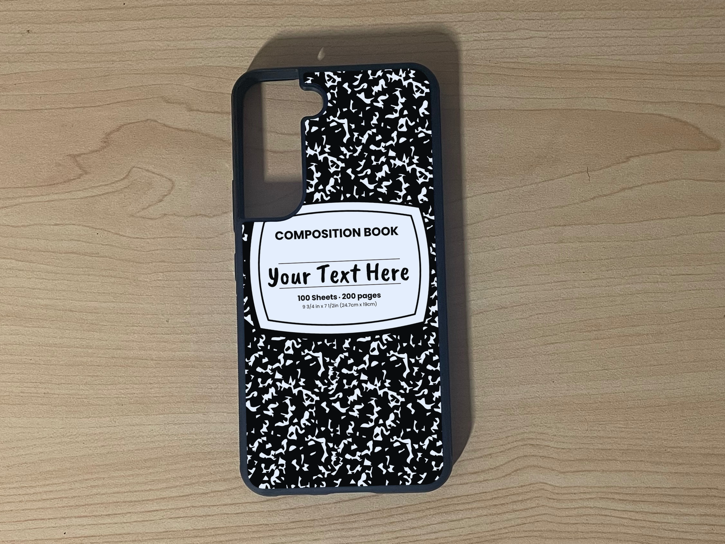 Custom phone case for the Samsung S21 and S21 Plus with a black and white composition notebook design with text "Your text here" on a wooden surface.