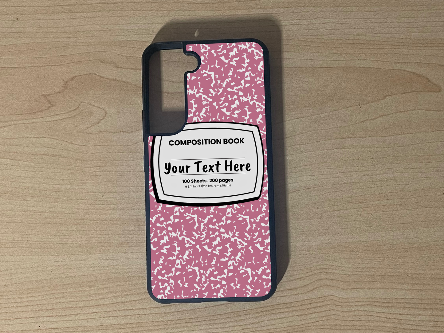 Custom phone case for the Samsung S21 and S21 Plus with a pink and white composition notebook design with text "Your text here" on a wooden surface.