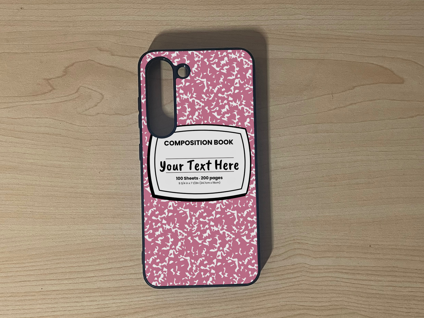 Custom phone case for the Samsung S23 and S23 Plus with a pink and white composition notebook design with text "Your text here" on a wooden surface.