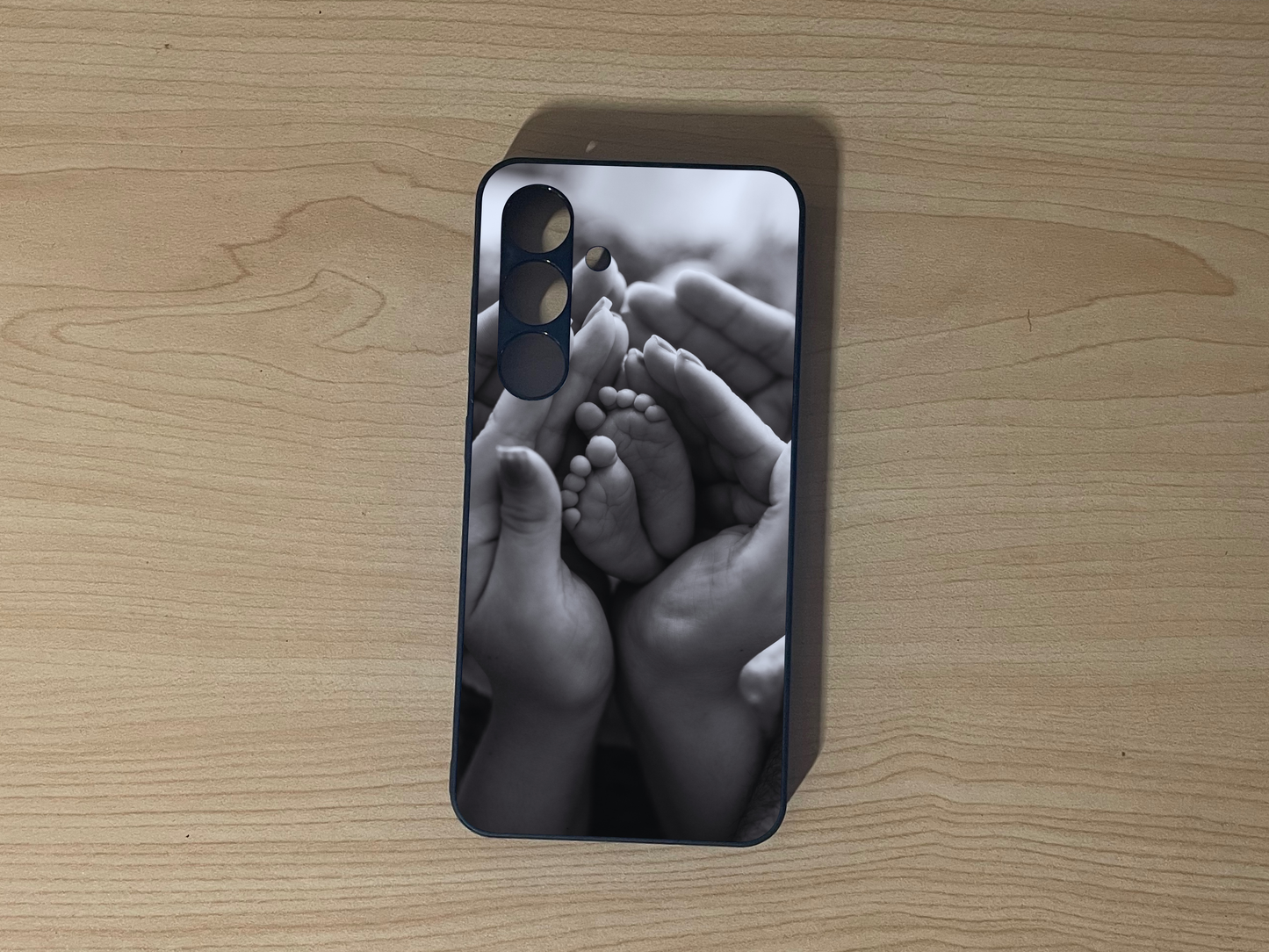 Custom phone case for the Samsung S24 and S24 Plus showing a black and white design of baby feet and hands on a wooden surface.