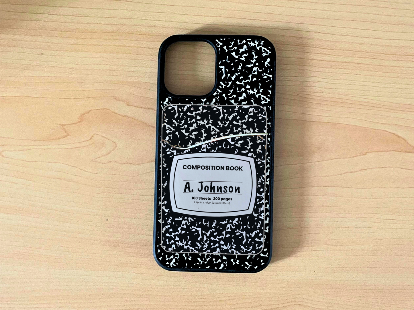 Phone case with black and white composition notebook design with matching adhesive card holder on a wooden background.