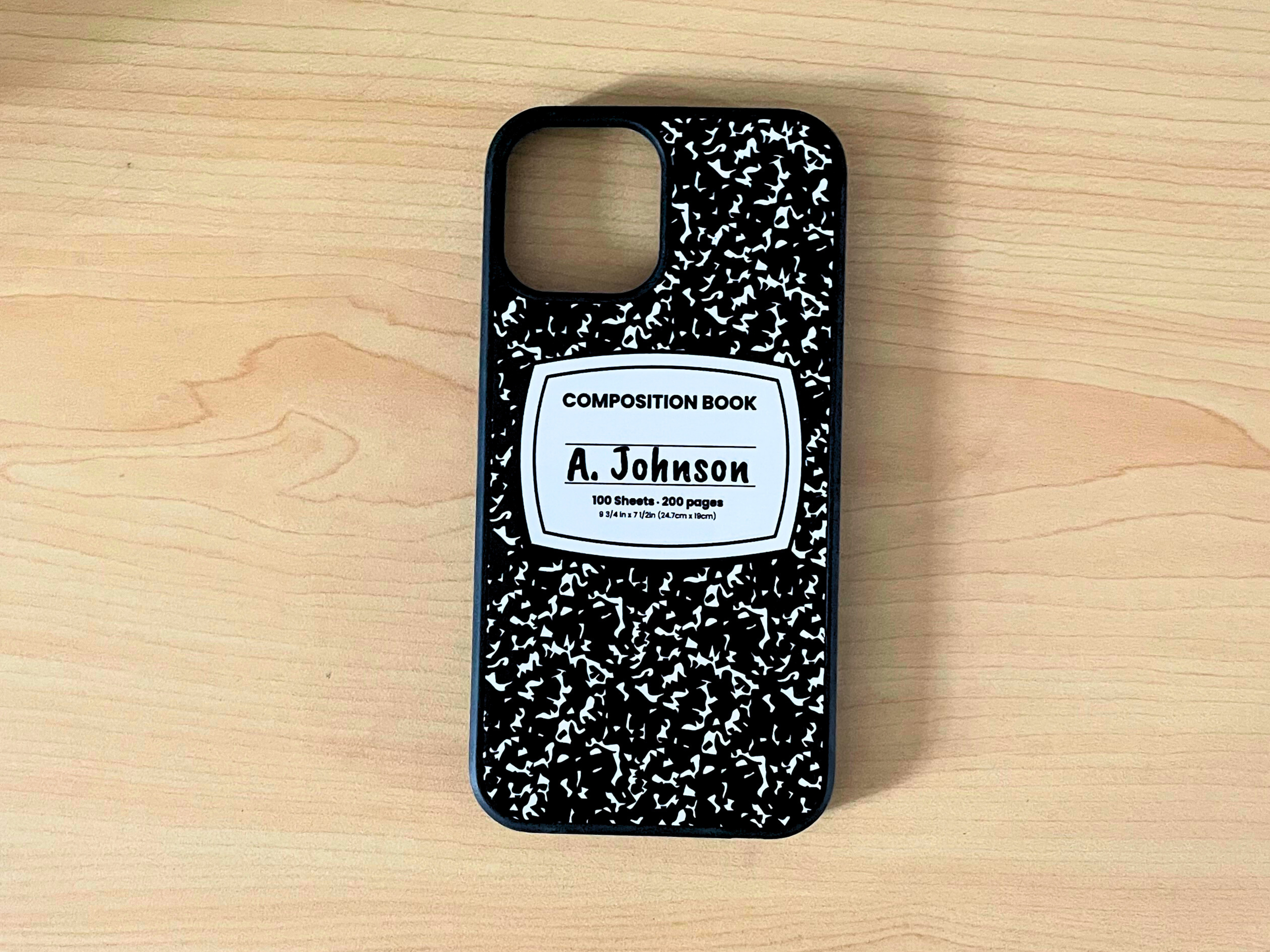 iPhone case with black and white composition notebook design and 'A. Johnson' written in the middle displayed on a wooden surface.
