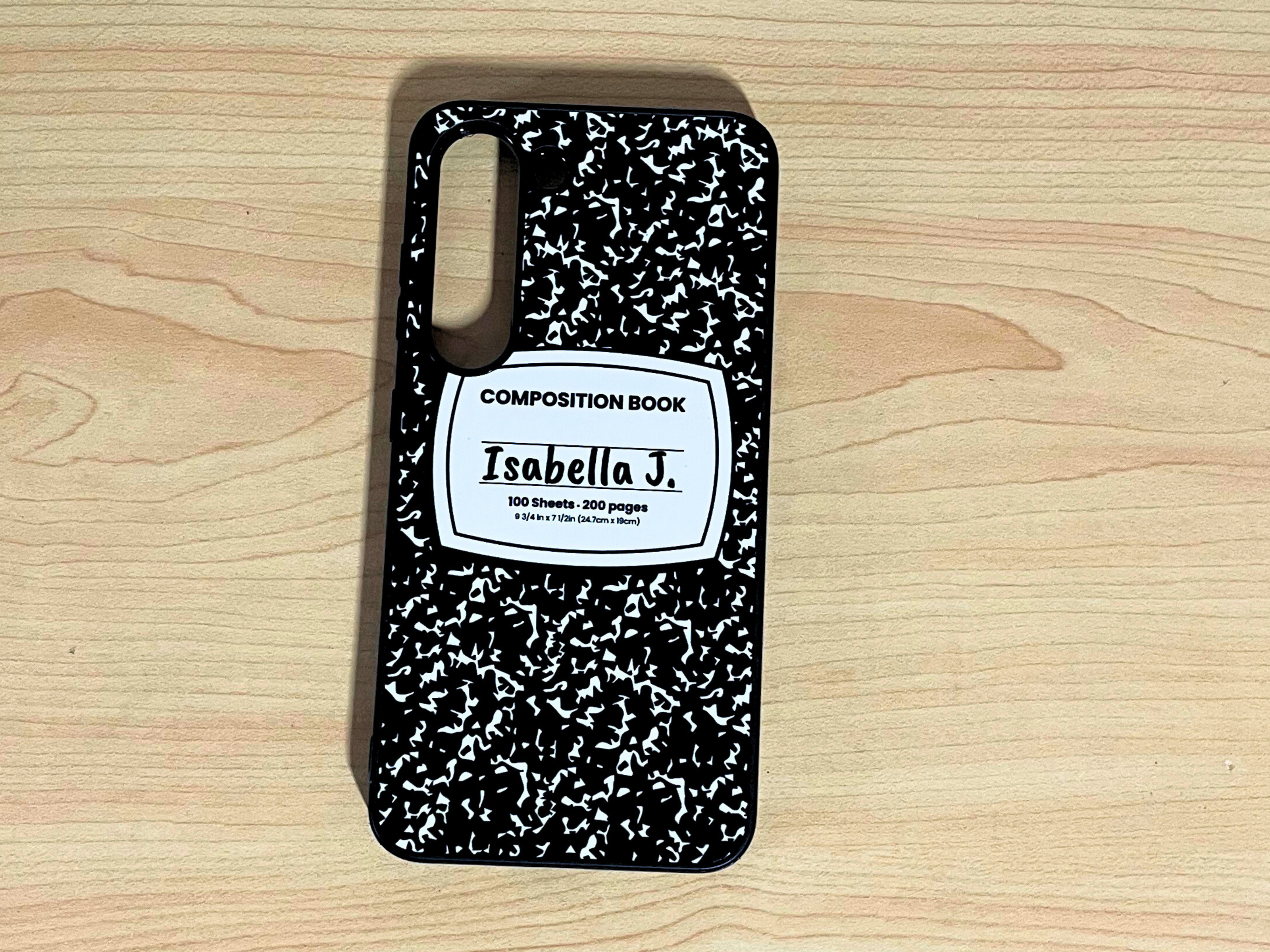 Samsung Galaxy case with black and white composition notebook design and 'Isabella J.' written in the middle displayed on a wooden surface.