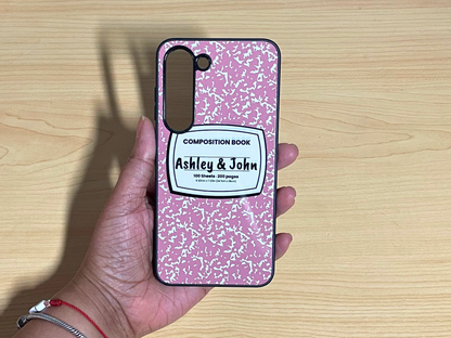 Samsung Galaxy case with pink and white composition notebook design and 'Ashley & John' written in the middle displayed on a wooden surface.