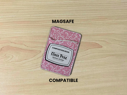 MagSafe compatible phone wallet/card holder with pink and white composition notebook design with customizable text "Class Prez" on the center on a wooden background.