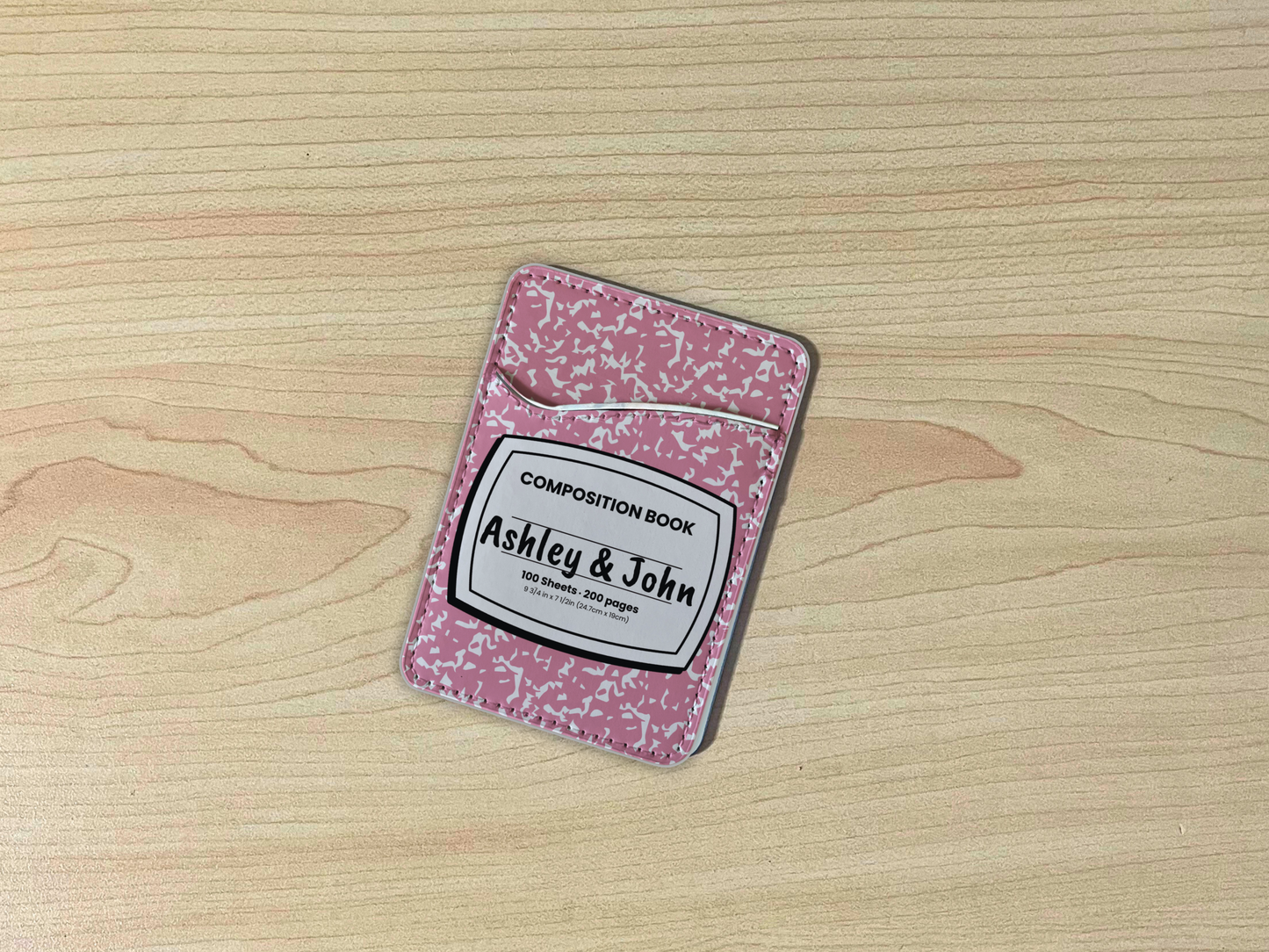 Pink and white composition notebook adhesive phone wallet/card holder with custom text "Ashley & John" in the center on a wooden surface