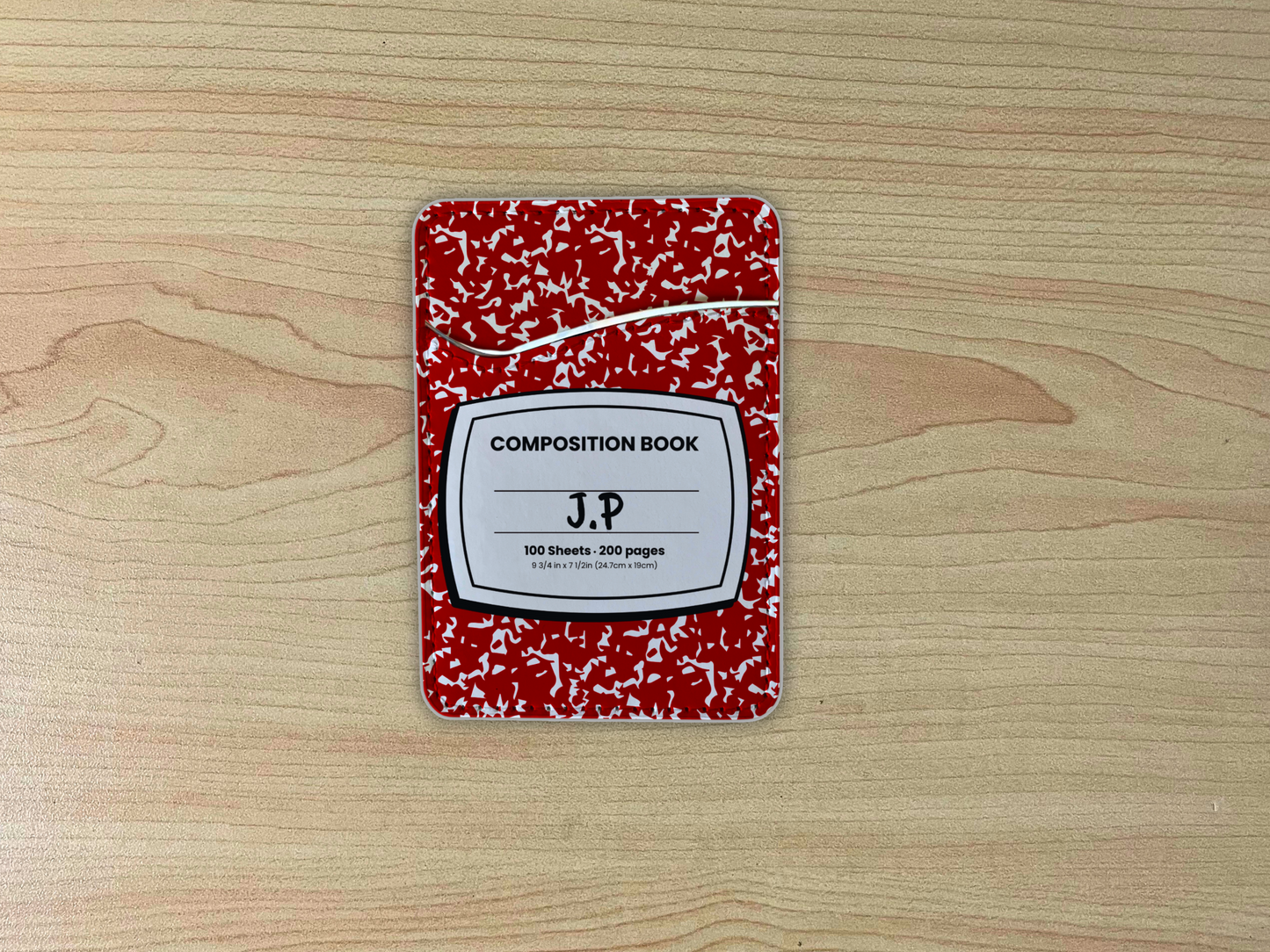 Red and white composition notebook adhesive phone wallet/card holder with custom text "J.P" in the center on a wooden surface