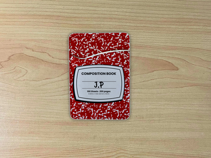 Red and white composition notebook adhesive phone wallet/card holder with custom text "J.P" in the center on a wooden surface