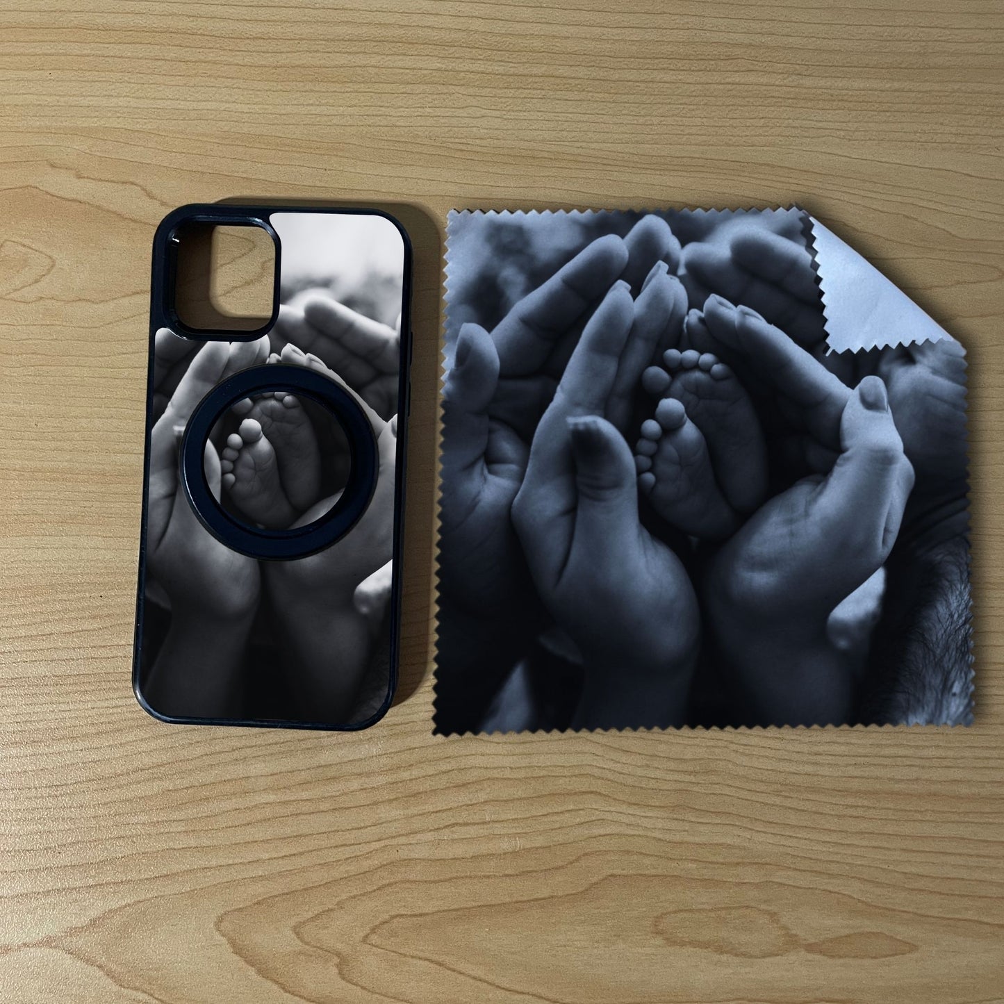 Custom MagSafe iPhone case bundle set with hands holding a baby's feet photo design with matching black phone grip and matching microfiber cloth phone screen cleaner on a wooden surface.