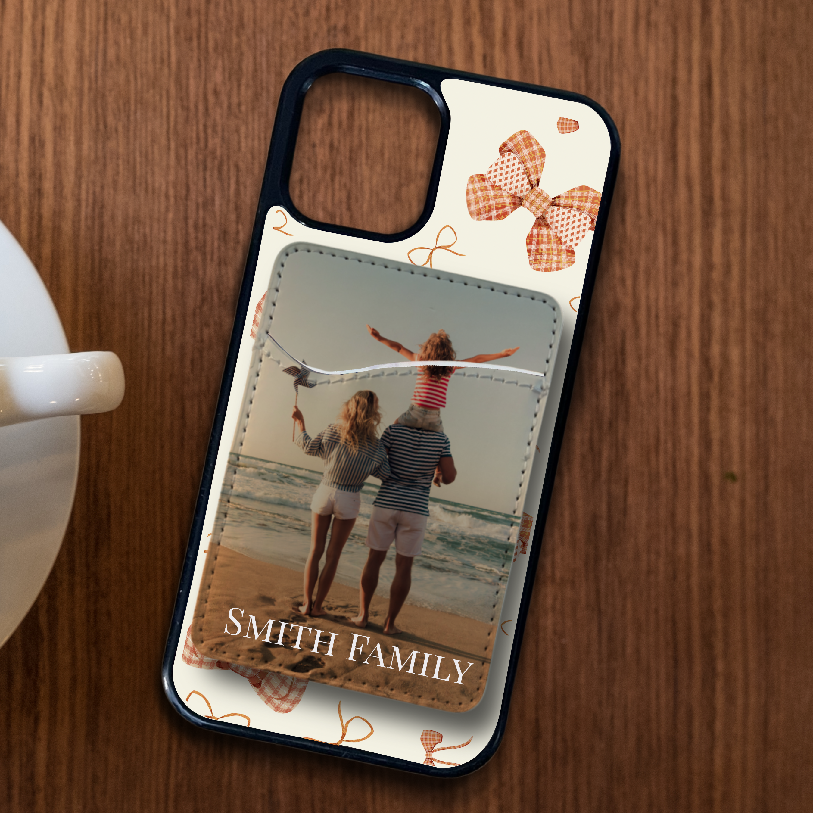 Custom phone case with a bow design on a sleek black background, featuring a personalized phone card holder attached to the back. The holder displays a vacation photo of a happy family with the text "Smith Family" printed below the image.