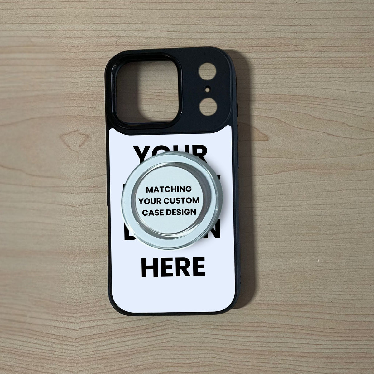 A MagSafe iPhone 17 Pro/17 Pro Max custom phone case with a black bumper with a clear MagSafe phone grip featuring placeholder text, resting on a light wood grain surface. 