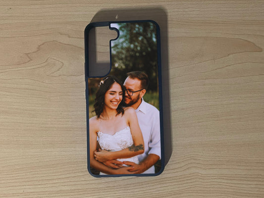 A custom protective Samsung Galaxy S 25 Ultra phone case with a printed photo of a smiling couple, displayed on a light wood grain surface.