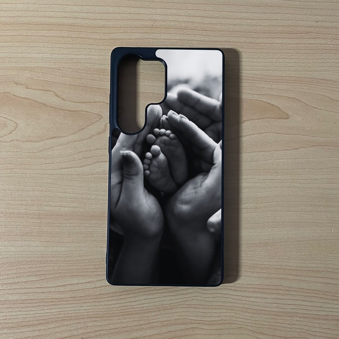Custom phone case for the Samsung S25 Ultra showing a black and white design of baby feet and hands on a wooden surface.